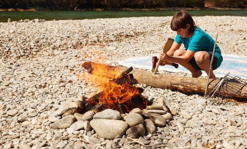 Mateo sitzt mit einem Beil am Feuer und bearbeitet ein Stück Holz