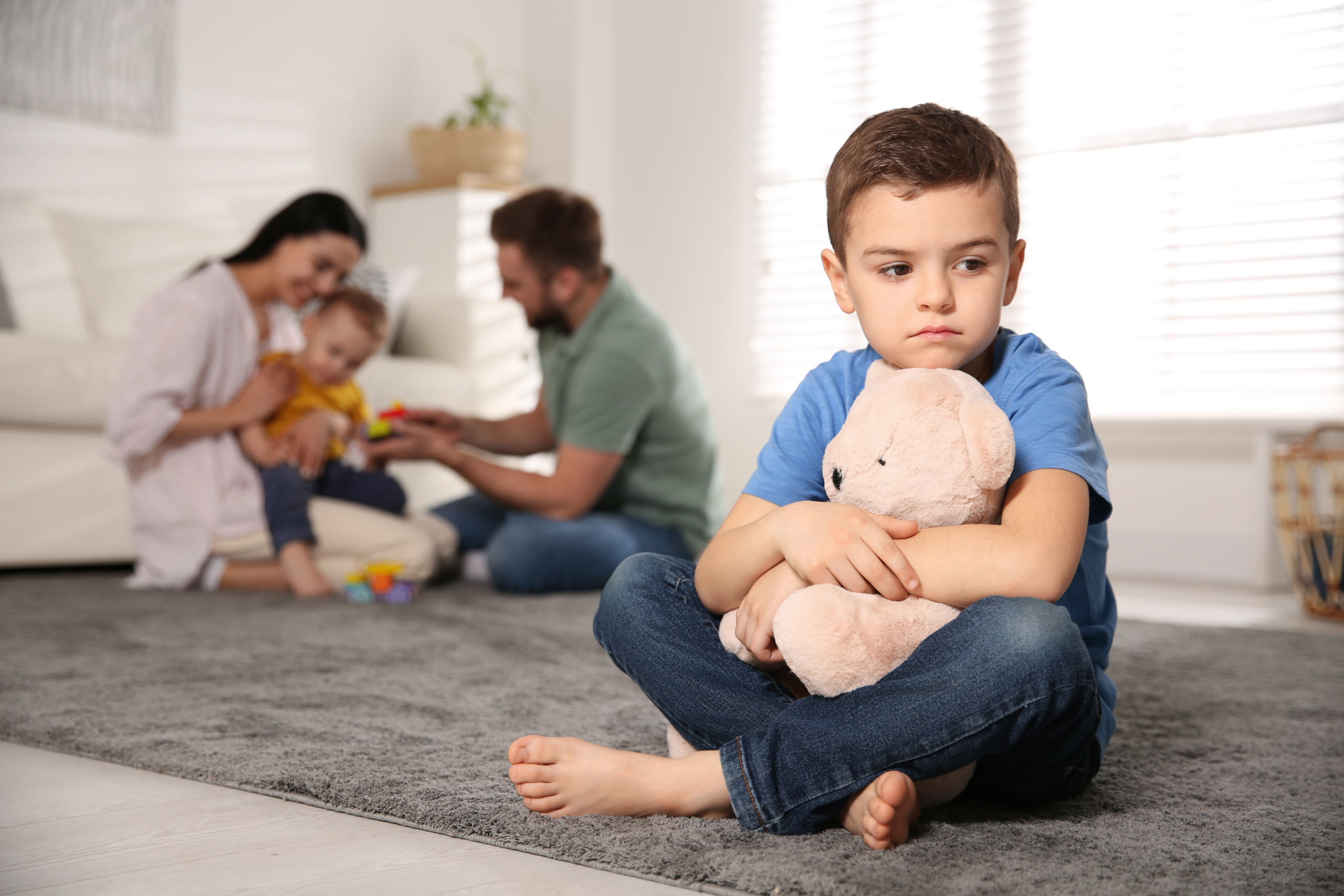Hänseler_Rescue Kids_Eifersucht_Advert_848x512px Ängstlicher Junge sitzt auf dem Teppich, Eltern mit kleinerem Geschwisterkind im Hintergrund